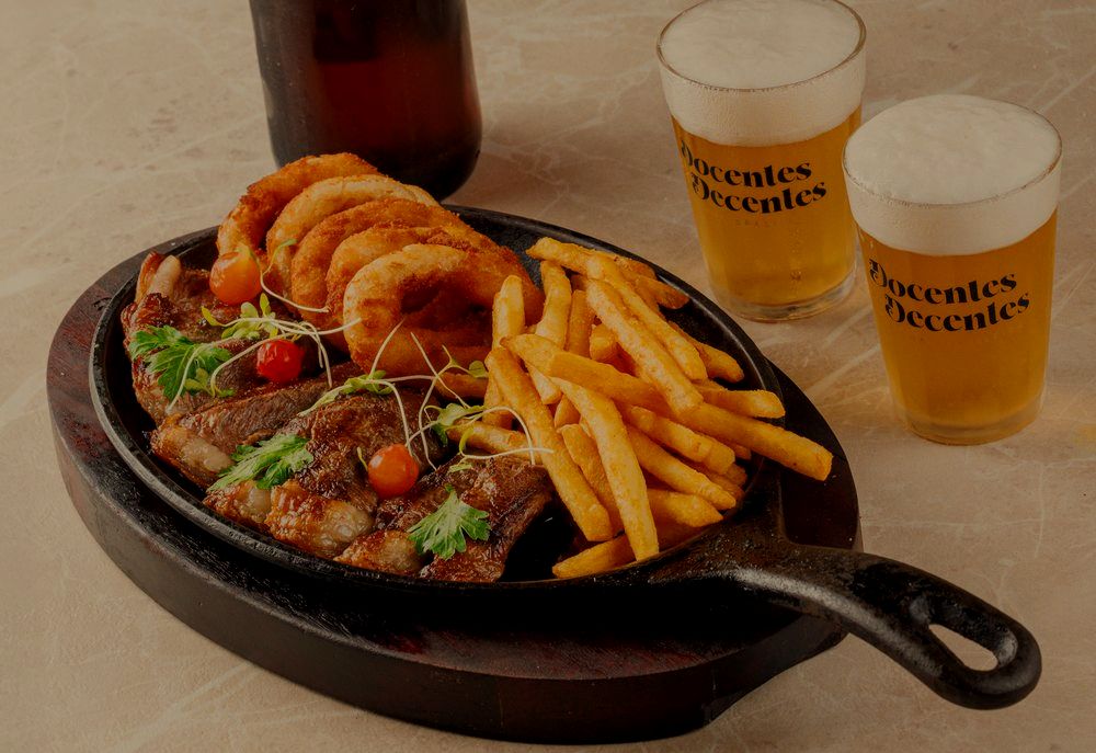 Chapa com carne, anéis de cebola e fritas, ao lado de copos com a marca da casa